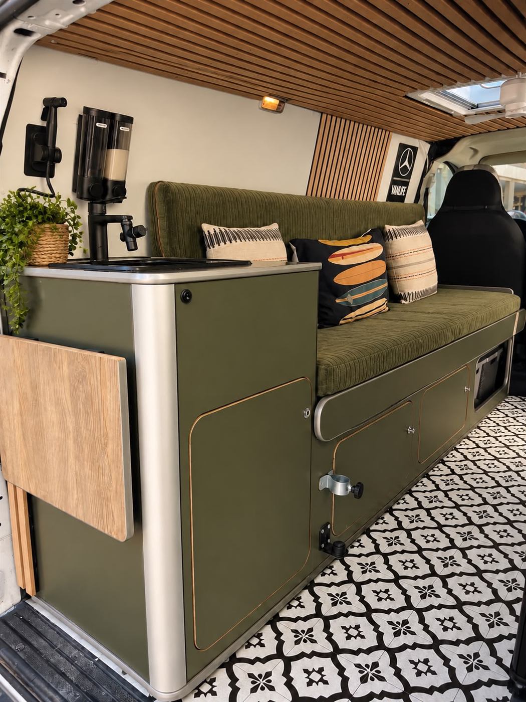 Mobiliario a medida verde oliva con fregadero, mesa abatible y banco-cama en una camper